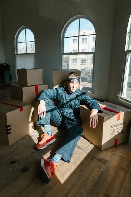 A young man wearing a blue work jumpsuit, red sneakers, and a patterned headband sits on the wooden floor inside a bright room with large arched windows, surrounded by several sealed cardboard moving boxes with red and black tape. The boxes vary in size and are stacked or positioned nearby, indicating a packing stage during a home relocation. Sunlight streams through the windows, illuminating the scene and casting shadows on the floor. The room appears to be part of a house in Kingston Vale, with a view of neighboring buildings outside. The image captures the process of packing and preparing belongings for a move, with Man and Van Kingston Vale providing professional removals and moving services, emphasizing organized and efficient packing and furniture transport.