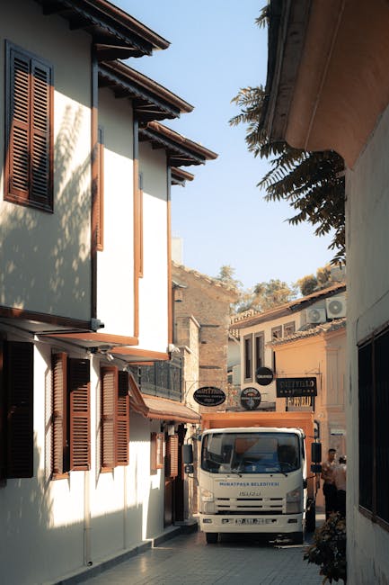 A narrow street lined with white buildings featuring dark wooden shutters, with a white van parked in the middle of the road. The van is part of a home relocation or moving process, with its front facing toward the camera. There are cardboard boxes, plastic wrapping, and blankets visible on or around the vehicle, indicating packing and transit preparations. An individual is standing nearby, possibly assisting with loading or unloading items. The scene is outdoors during daylight, with a clear sky and some foliage visible in the background, capturing the tight urban environment typical for furniture transport and house removals. The setting is consistent with professional removals services offered by Man and Van Kingston Vale, emphasizing the logistical aspect of home relocations within a residential area.