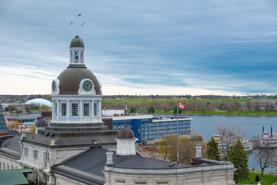 Aerial view of a cityscape showing a prominent historic building with a large domed clock tower, white walls, and dark roofing, situated near a river. Surrounding the building are other structures including modern and traditional buildings, with some featuring dark roofing and brick facades. In the background, a blue multi-storey building is visible along the riverbank, alongside trees with sparse foliage, indicating early spring. A Canadian flag is flying on a flagpole near the water, while boats are docked along the river's edge. The sky is partly cloudy with patches of blue, and the scene is illuminated by natural daylight. This setting relates to house removals and home relocation activities, illustrating the importance of careful packing, loading, and transport of furniture and boxes for efficient moving services, as provided by Man and Van Kingston Vale.