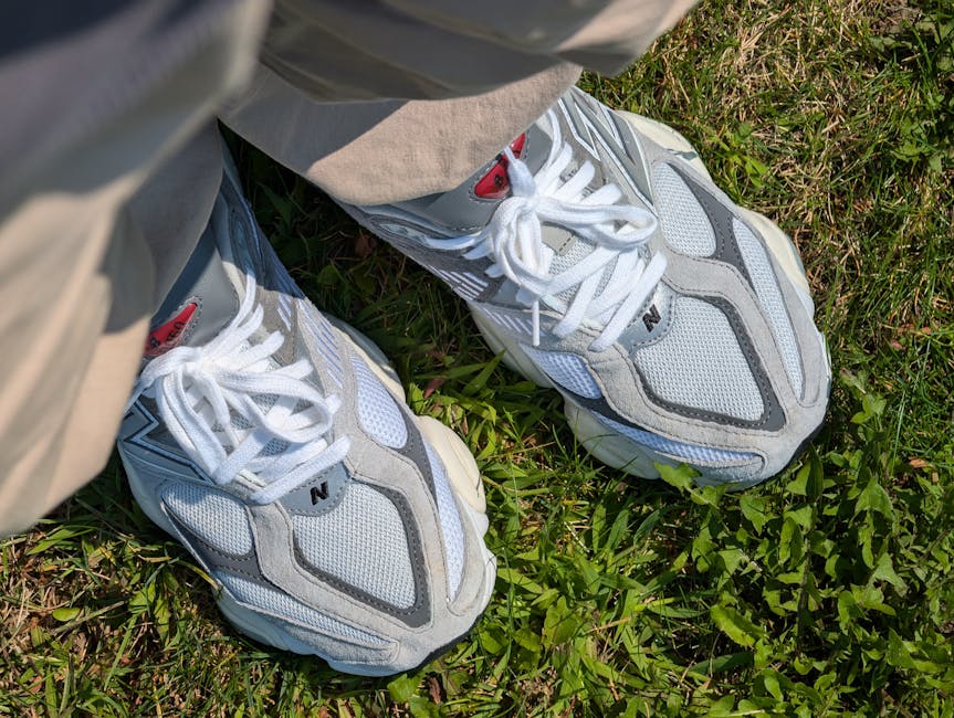 A close-up view of a person's feet wearing white, grey, and black athletic sneakers with white laces, standing on a patch of green grass with small plants. The individual is dressed in beige pants, and the photograph appears to be taken outdoors during daylight. The image also shows part of the person's beige jacket or coat. In the background, a moving box or appliance may be partially visible indoors or in a doorway, indicating a home relocation or packing process. The setting suggests preparation for a move or loading an item, with the focus on the footwear and ground. This scene is relevant to house removals and furniture transport, with natural lighting emphasizing the details of the sneakers and grassy surface, consistent with a professional storage or moving service like Man and Van Kingston Vale.