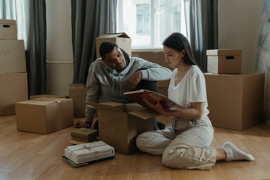 A man and a woman are inside a residential property during a home relocation process, surrounded by packed cardboard boxes of various sizes, some labeled with handwritten notes such as 'books'. The woman, sitting on the wooden floor, is reading a book while the man, leaning slightly forward, attentively looks at her and the nearby boxes. The boxes are arranged around them, with some stacked near a window with open curtains, indicating natural light illuminating the room. Visible items include a cardboard box with a label reading 'books', a smaller box wrapped with string, and an open box with packing paper. The scene captures the packing stage of a furniture transport and moving process as part of house removals, with the environment suggesting an organized moving preparation within the home from Man and Van Kingston Vale.
