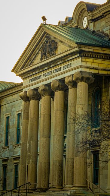 Exterior view of Frontline County Court House showing its classical architectural style with large stone columns supporting a triangular pediment, which features an ornate carved emblem. The building's facade is made of light-colored stone, with rectangular windows, some of which are arched at the top. In the foreground, there are several cardboard moving boxes stacked on a paved pavement, with a black plastic-wrapped item and a moving trolley nearby. The scene depicts a home relocation or furniture transport process outside the court building, indicating preparations for a house move or legal proceedings related to property. The overall lighting suggests daytime with natural sunlight illuminating the structure and the moving materials.
