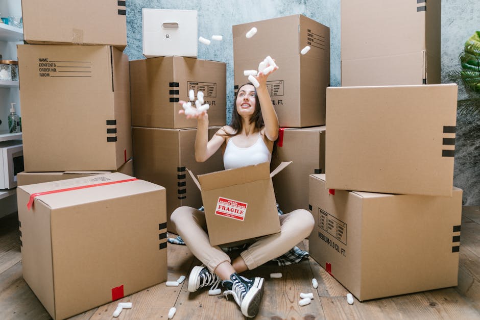 A young woman sitting on the floor inside a residential property, surrounded by several large cardboard moving boxes of various sizes, some labeled with packing details. She is wearing casual clothing and sneakers, and is in the process of unpacking or packing, as she is tossing packing foam into the air. A cardboard box with a red 'Fragile' label is positioned in her lap, and additional packing materials are scattered around her. The background features a textured wall, a potted plant, and a doorframe, indicating an indoor setting consistent with a home environment undergoing a furniture transport and home relocation process. The scene captures the active stage of packing and loading routines typical in house removals, with natural lighting emphasizing the practical aspects of moving logistics. Man and Van Kingston Vale’s professional removals service may assist with this home moving activity.
