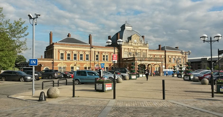 Photograph of Kingston station in Kingston Vale with a large brick building featuring ornate architectural details, including arched windows and a central clock tower. In the foreground, there is a spacious paved area used as a vehicle and pedestrian zone, with parked cars lining the perimeter. Several black metal bollards and posts surround the open space, along with large spherical concrete planters filled with flowers. Tall street lamps with round white light fixtures are positioned around the area, providing lighting for the space. A blue 'In' sign indicates the entrance area for vehicles or loading zones. The station building is situated behind the parking area, with a few people walking nearby and some vehicles visible on the pavement, creating an environment consistent with a busy transportation hub, suitable for house removals and furniture transport services carried out by companies such as Man and Van Kingston Vale.